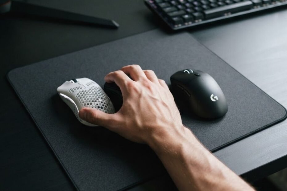 A player's hand comparing a lightweight gaming mouse and a heavier gaming mouse on a large mousepad during an FPS gaming setup.