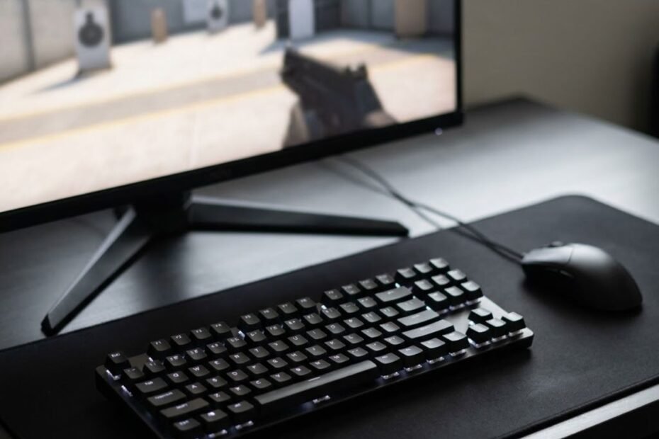 A gaming keyboard and mouse on a desk beside a monitor showing a first-person shooter training environment